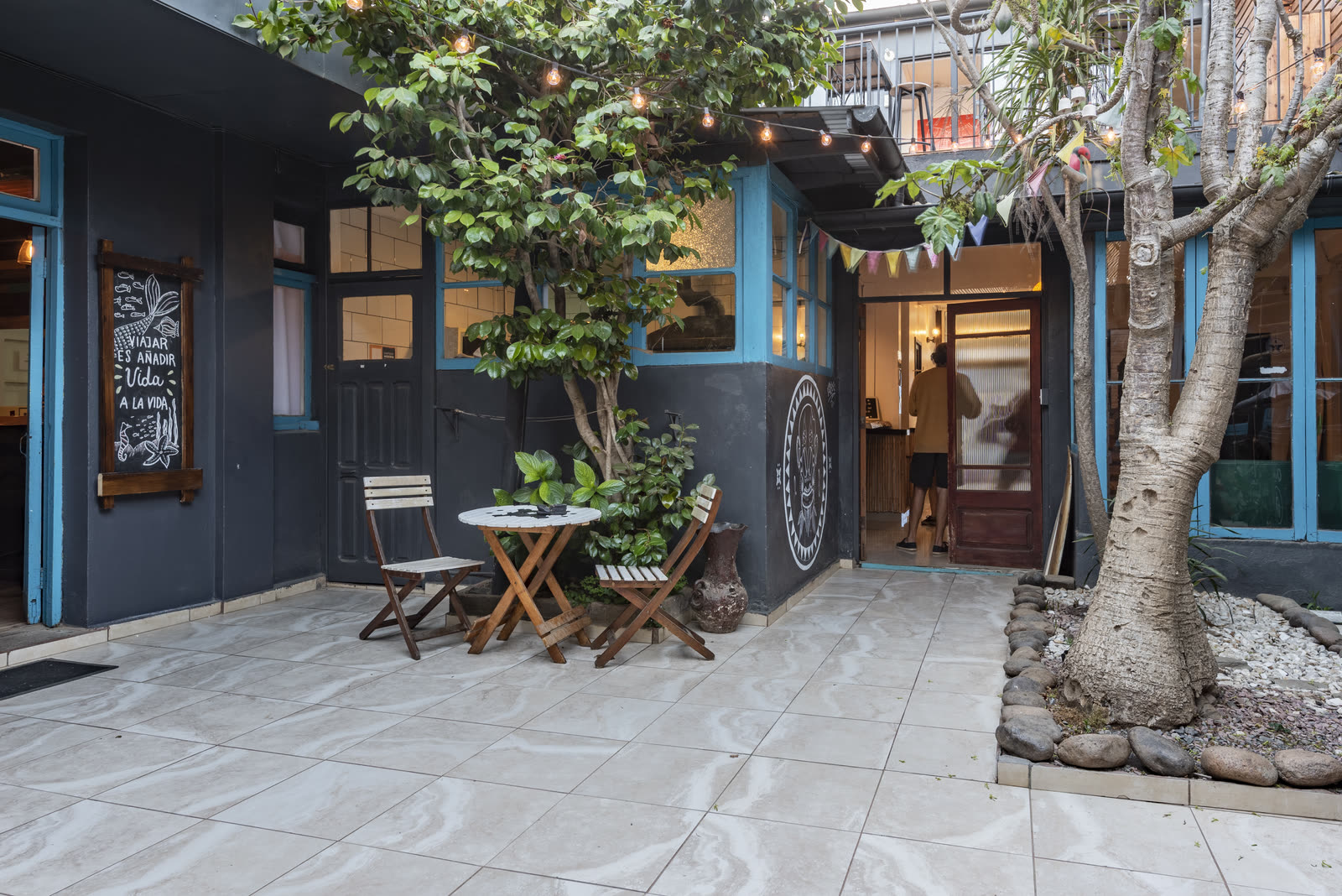Patio interior con un árbol grande, muros azules y una mesa con sillas